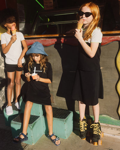 Young girl wearing black skirt and two tone tee, wearing roller skates and eating ice cream, another girl sitting eating ice cream and a boy standing and eating ice cream