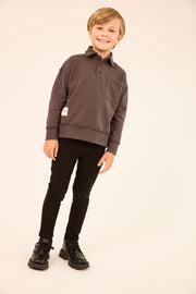 A young boy modeling a dark brown long-sleeve polo sweatshirt, paired with black pants and black boots, standing against a plain off-white studio background.