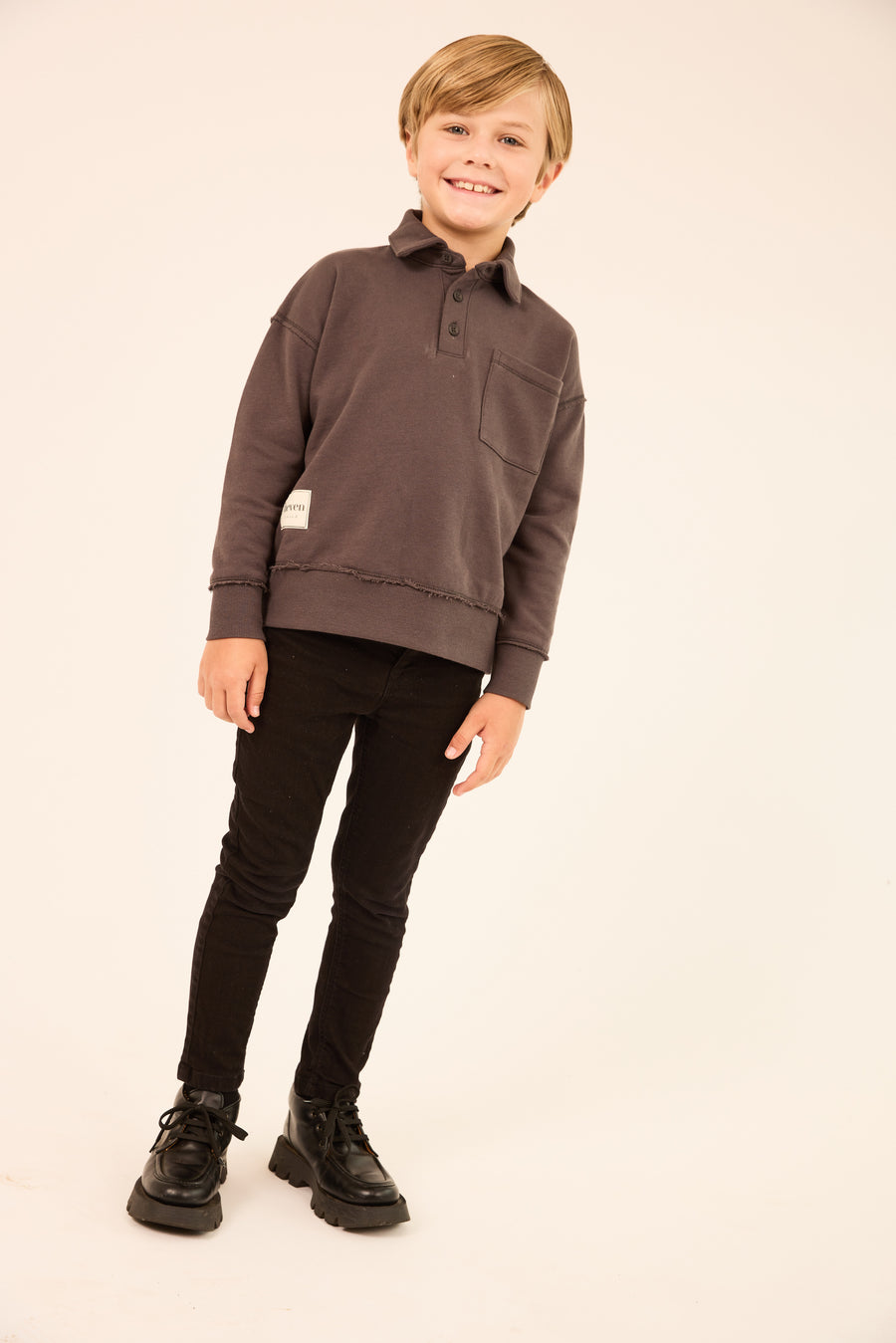A young boy modeling a dark brown long-sleeve polo sweatshirt, paired with black pants and black boots, standing against a plain off-white studio background.