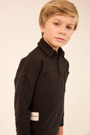 A young boy with blonde hair wearing a dark brown long-sleeve polo shirt, photographed against a plain light beige background.