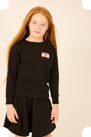 A young girl with long red hair wearing a black long-sleeve sweatshirt and matching black skirt, featuring a small red and white 'SWEATSHIRTS' patch on the left chest, set against a light beige background with white circular bokeh overlays.