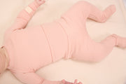 A baby lying on their back wearing a light pink footed sleeper on a white blanket.