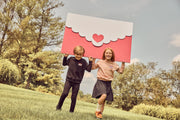 Two children running outdoors on a grassy hill holding a large red and white envelope-shaped sign with a small red heart in the center. Trees and blue sky are visible in the background.
