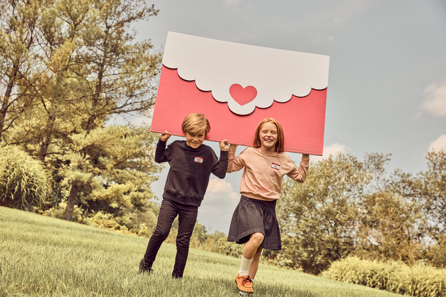 Two children running outdoors on a grassy hill holding a large red and white envelope-shaped sign with a small red heart in the center. Trees and blue sky are visible in the background.