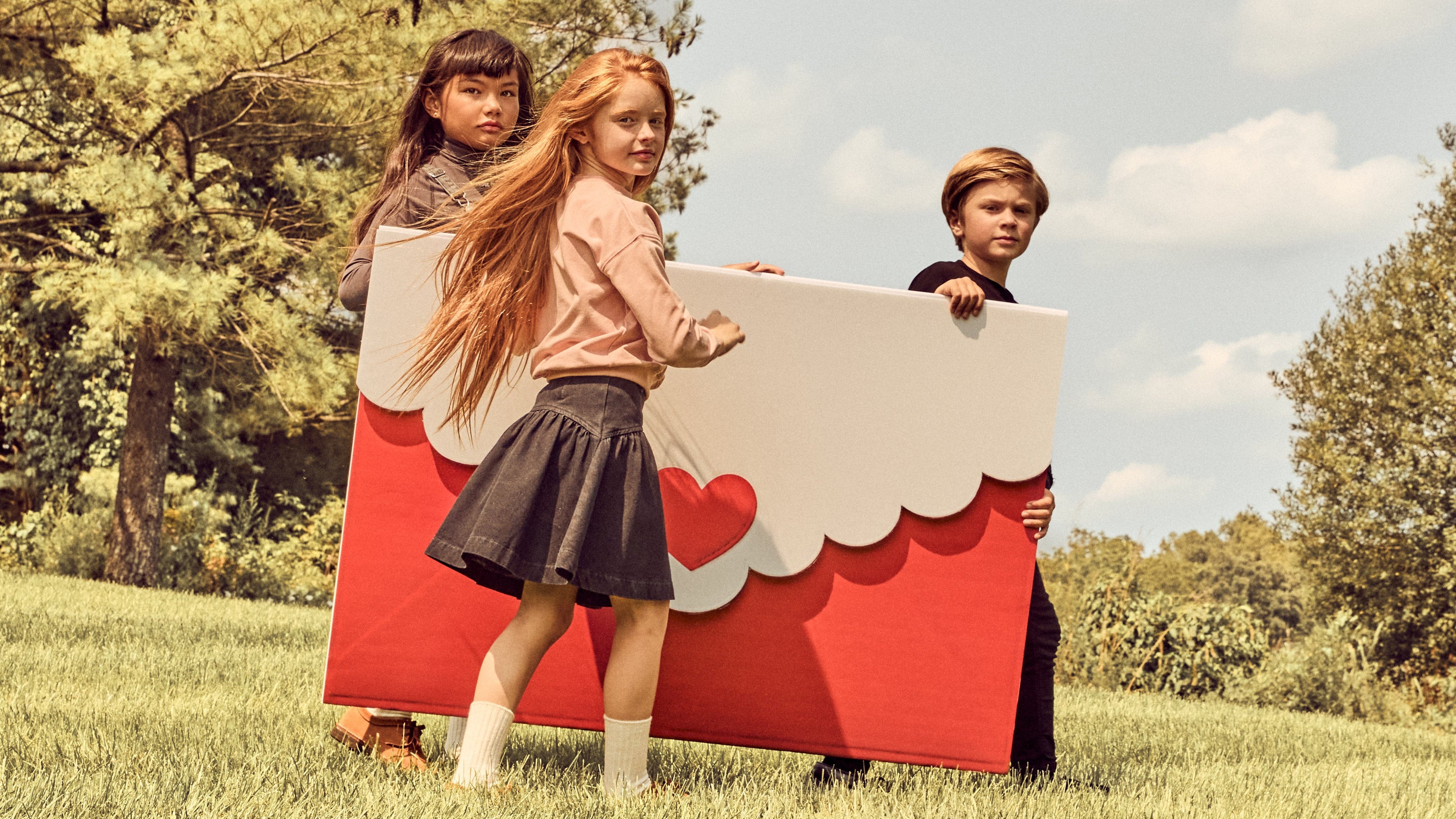 Three children outdoors on a grassy lawn under a cloudy sky, holding a large red and white cardboard box with scalloped edges and heart details.