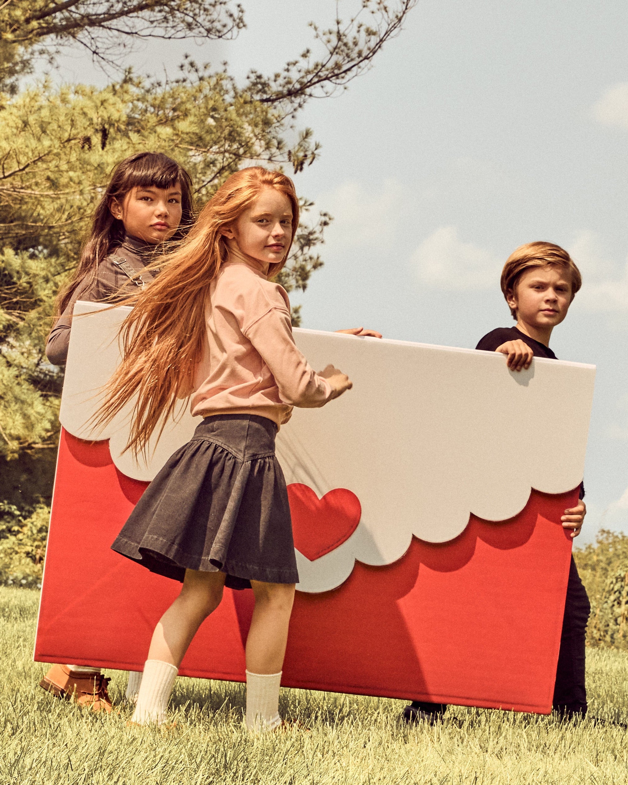 Three children outdoors on a grassy lawn under a cloudy sky, holding a large red and white cardboard box with scalloped edges and heart details.