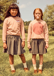 Two young girls standing outdoors in a grassy field, wearing matching outfits consisting of dusty pink long-sleeve tops and dark gray pleated skirts.