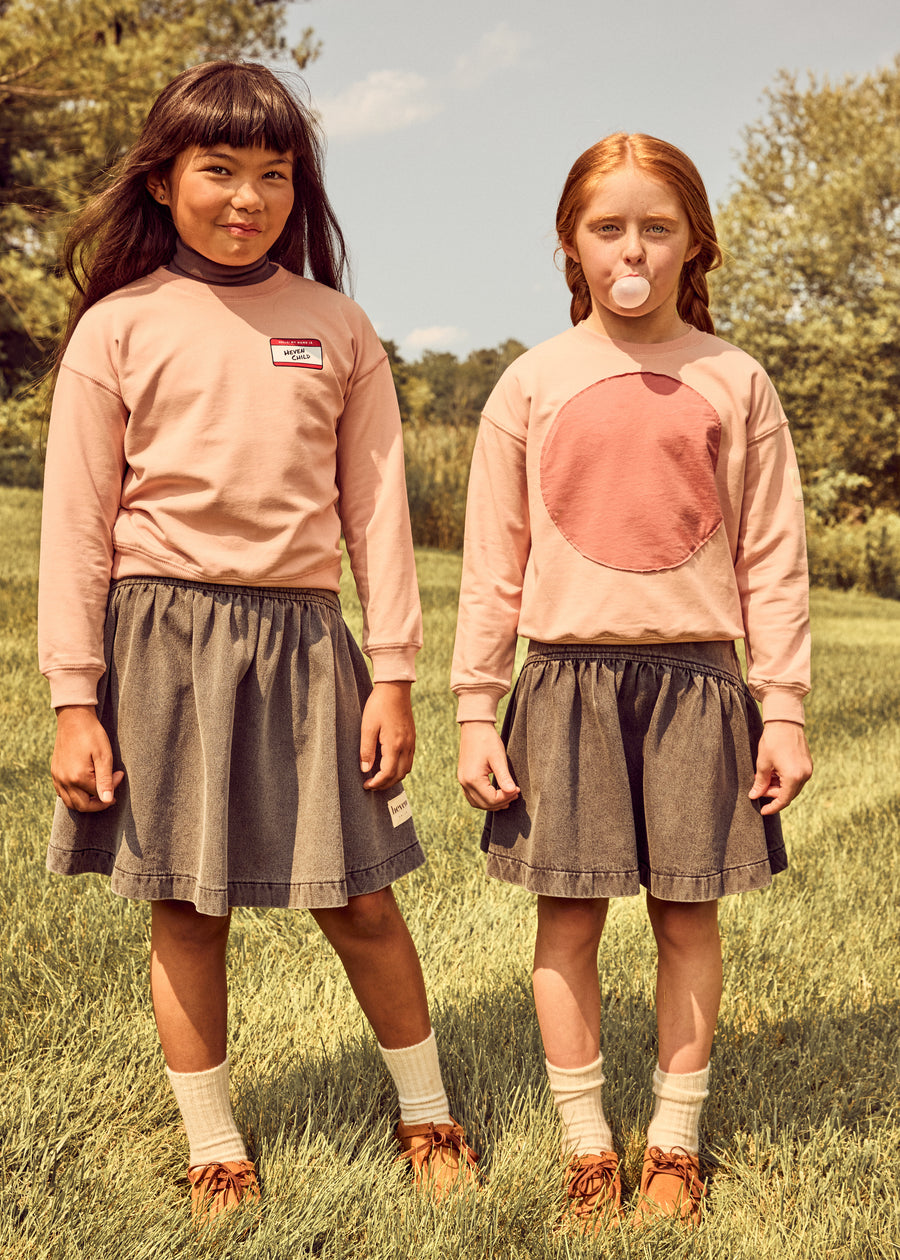 Two young girls standing outdoors in a grassy field, wearing matching outfits consisting of dusty pink long-sleeve tops and dark gray pleated skirts.
