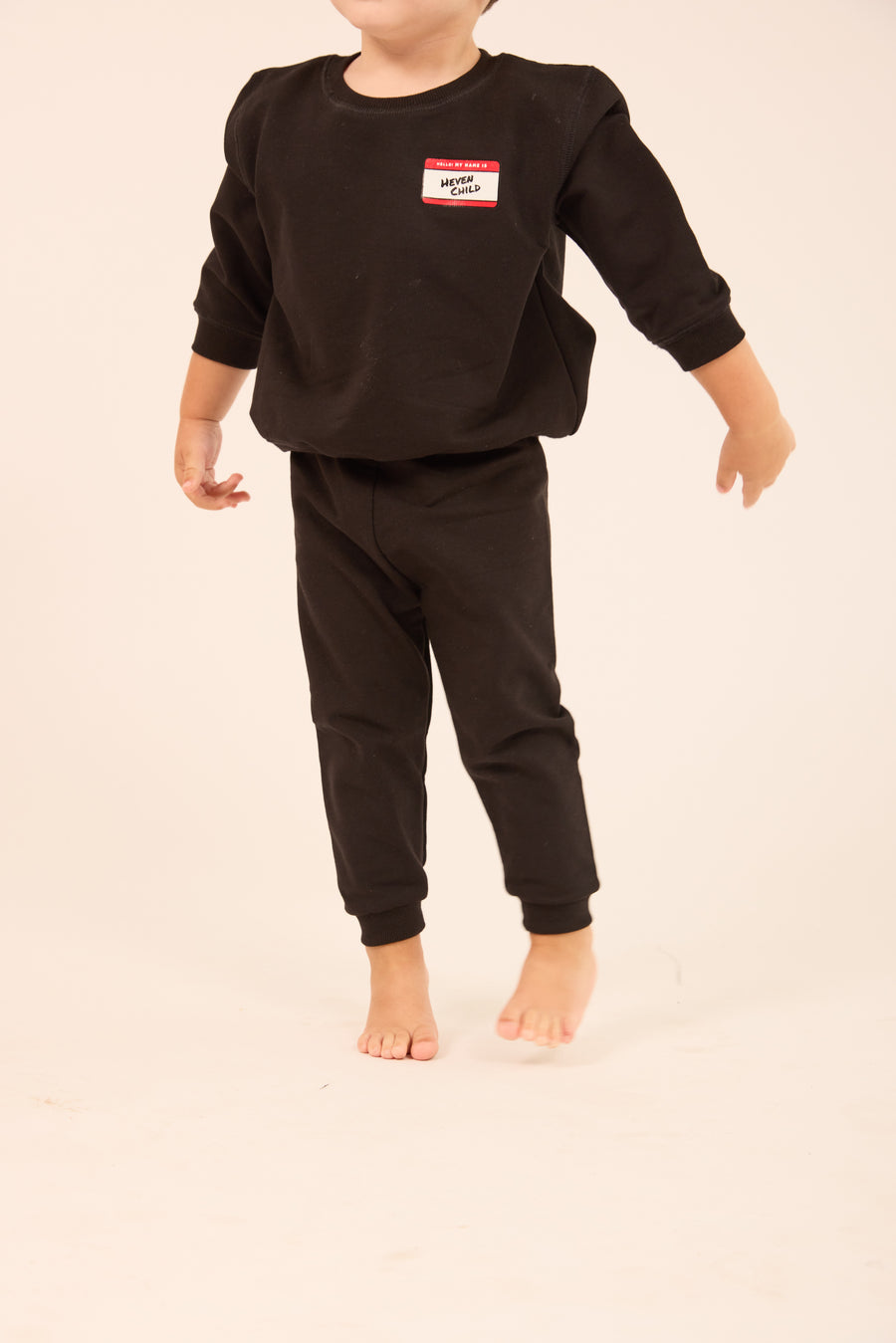 A toddler wearing a black long-sleeve sweatshirt and matching black pants, standing barefoot against a plain light beige studio background. The sweatshirt has a small red, white, and black name-tag style graphic on the left chest.