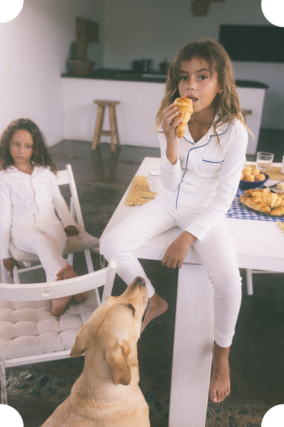 Young girl sitting on table wearing white pajamas with a dog looking at her and another girl in the background. 