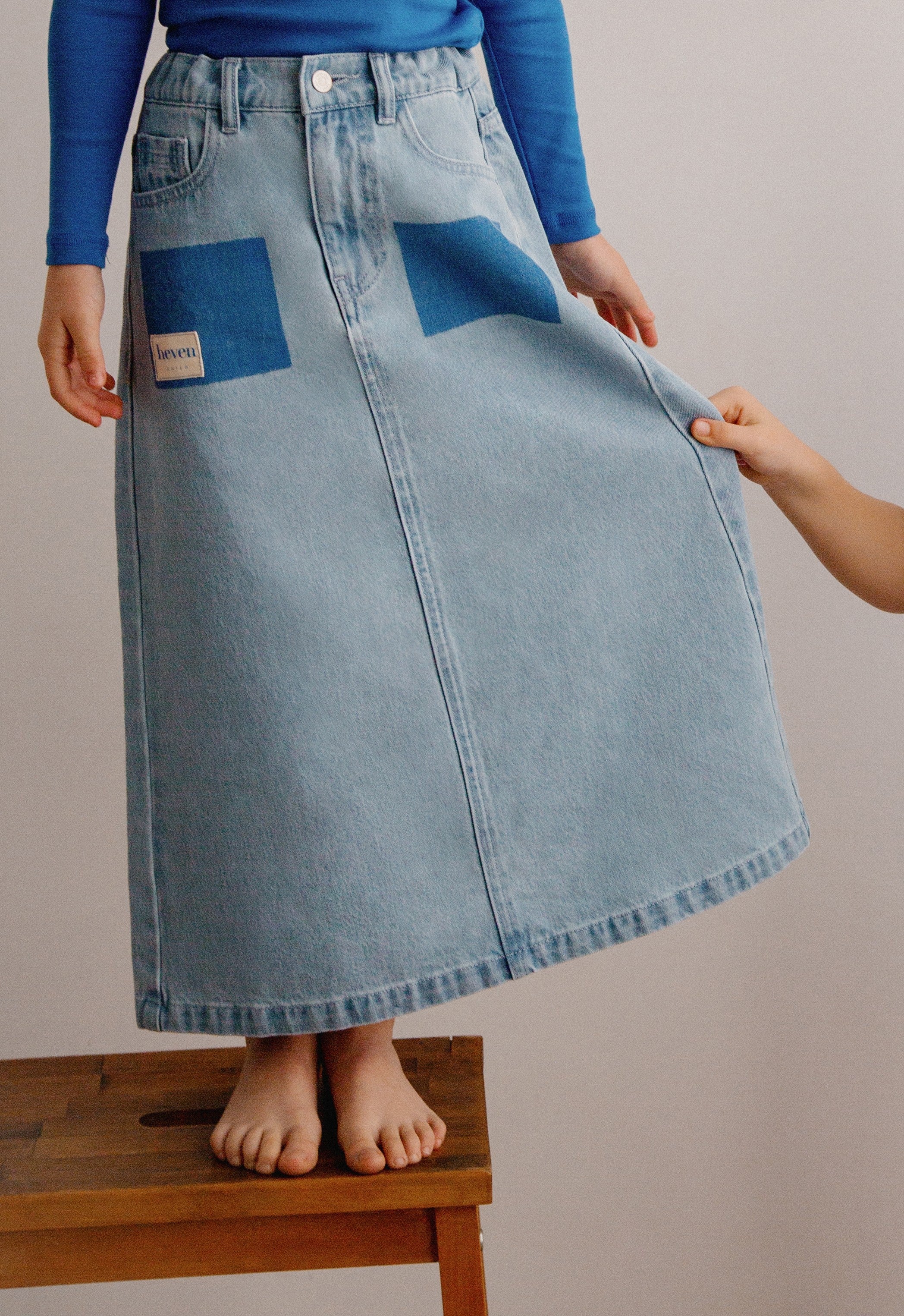 Person wearing a blue denim skirt with a visible brand label, standing on a wooden stool against a plain background.