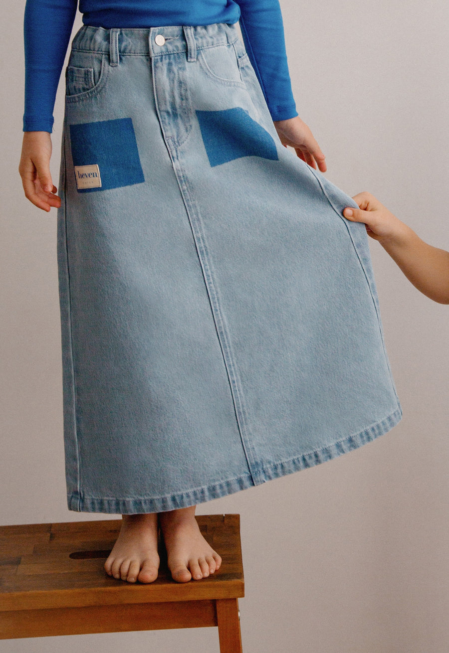 Person wearing a blue denim skirt with a visible brand label, standing on a wooden stool against a plain background.