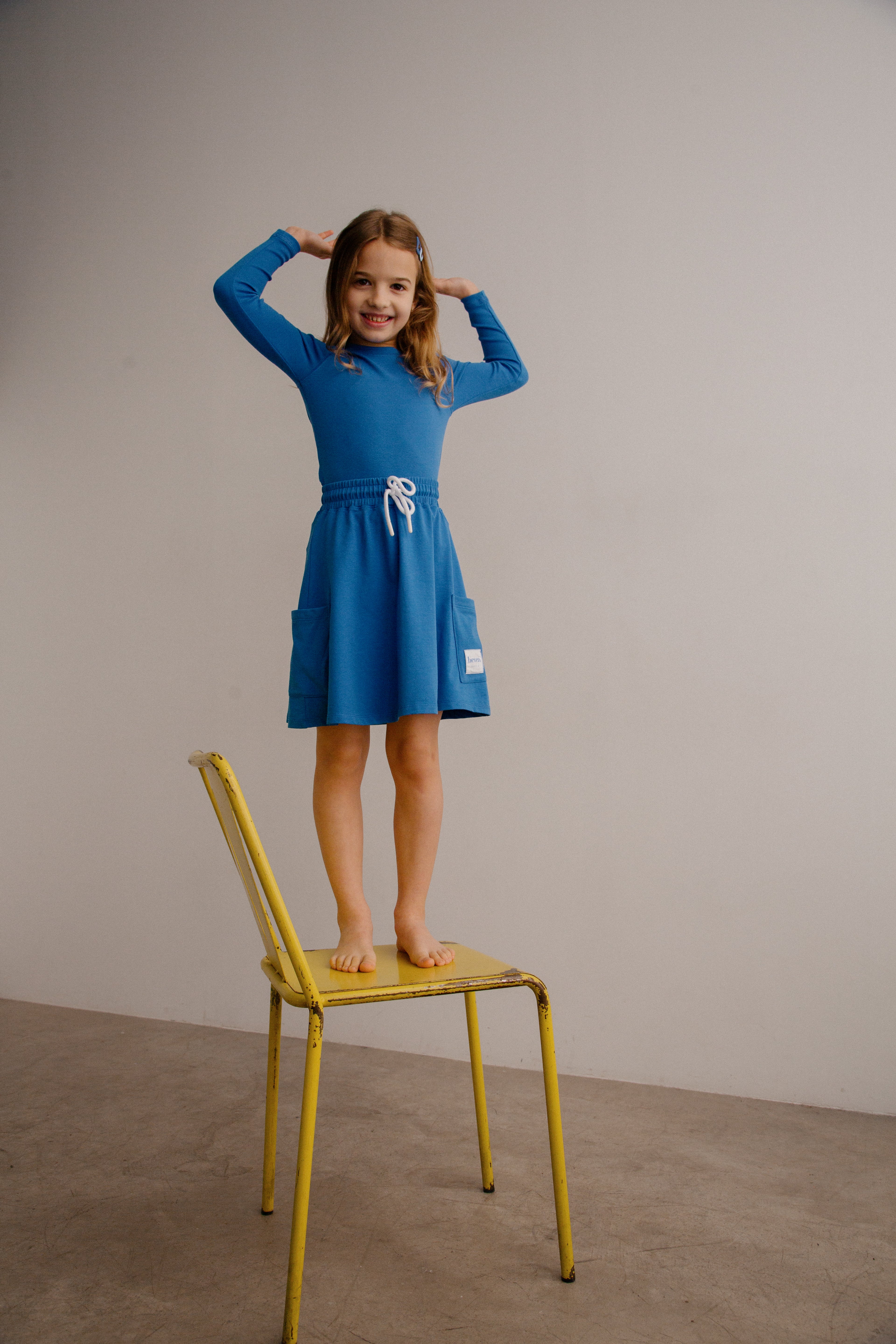Young girl in a blue dress standing on a yellow chair against a plain wall.