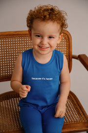 Child wearing a blue sleeveless shirt with text, sitting on a wooden chair.
