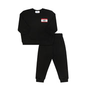 A two-piece kids' sweatshirt and jogger pants set in black. The sweatshirt features a small rectangular chest patch with a red top border, white background, and black text reading 'MONTI CROWE'.