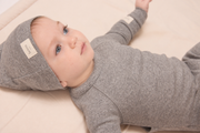 A baby lying on a cream-colored blanket, wearing a matching gray knit one-piece outfit with a hooded hat. The hat features a small white fabric label with the brand name 'lelequy' visible.