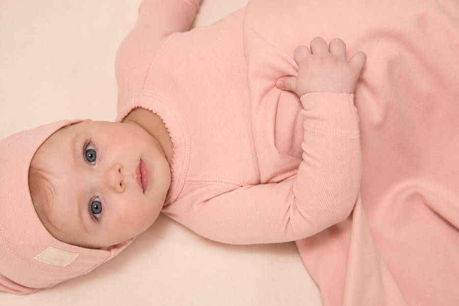 A baby wearing a light pink long-sleeve onesie and matching hat, lying on a soft cream-colored blanket.