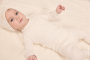 A baby lying on a soft cream-colored blanket, wearing a matching long-sleeve footed onesie and bonnet in a warm off-white or cream color.