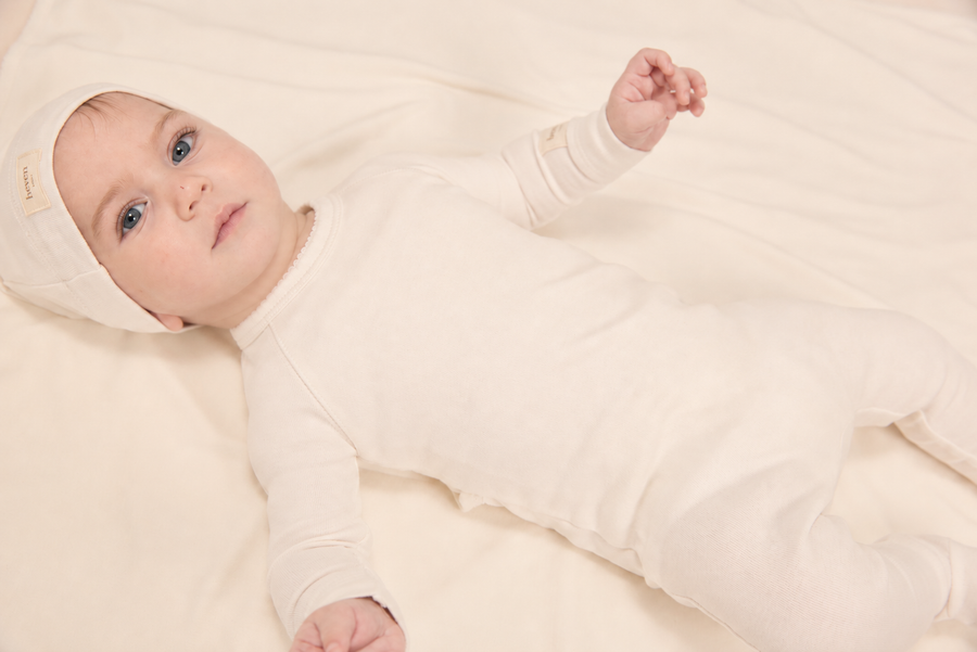 A baby lying on a soft cream-colored blanket, wearing a matching long-sleeve footed onesie and bonnet in a warm off-white or cream color.