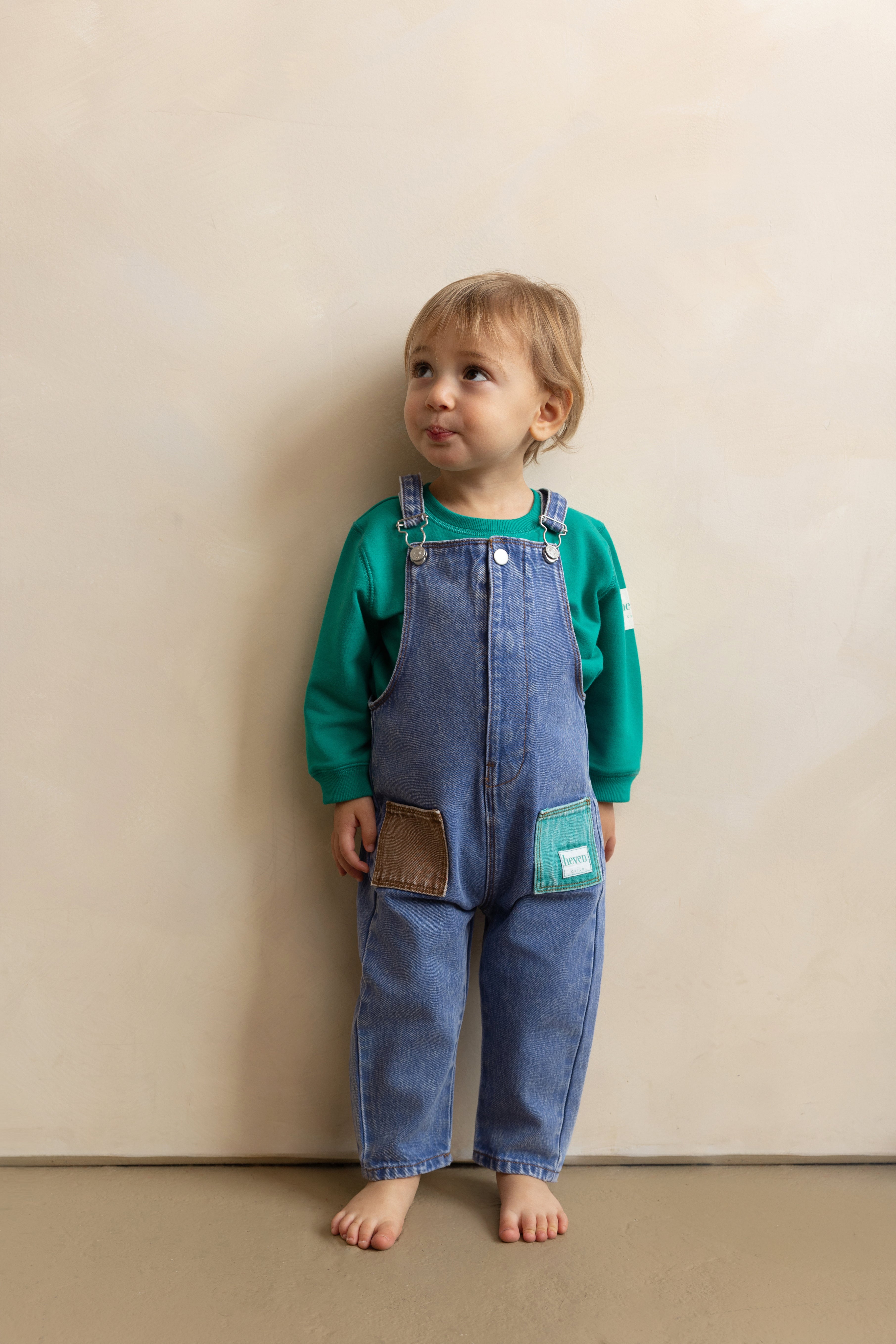 Child wearing blue denim overalls and a green shirt against a beige wall.