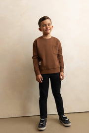 Child wearing a brown sweatshirt and black pants standing against a beige wall.