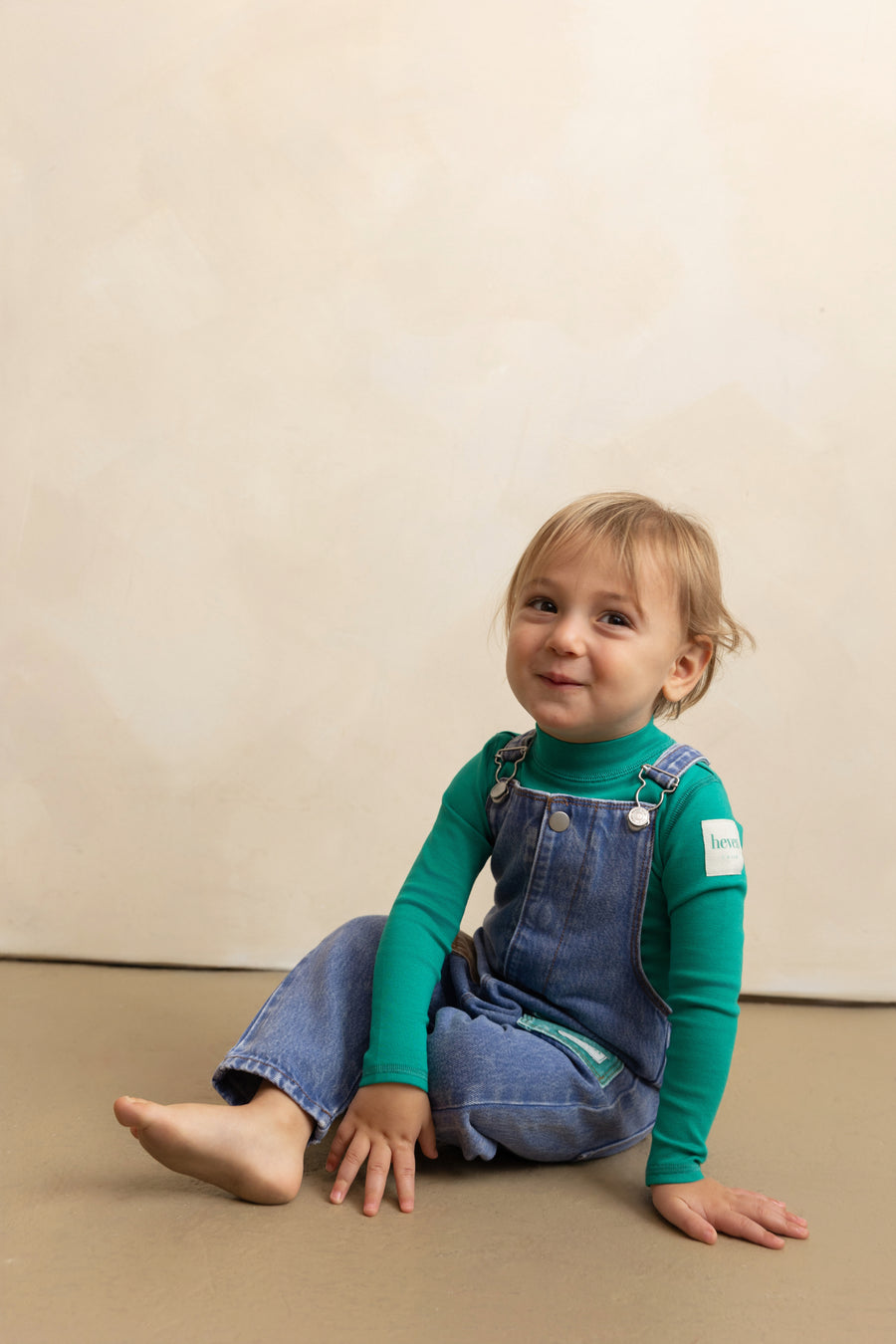 Child wearing a green long-sleeve shirt and blue denim overalls sitting on a beige floor.