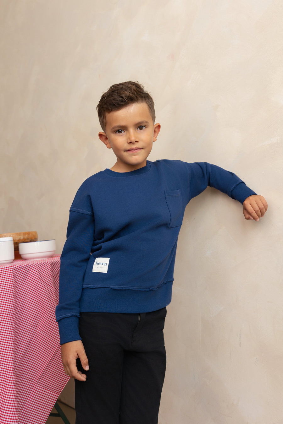 Child wearing a blue sweatshirt with a visible brand logo, standing against a beige wall.