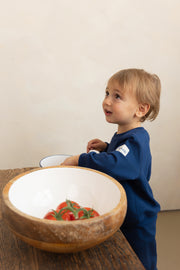 Child in a blue outfit standing next to a wooden bowl with tomatoes on a wooden table.