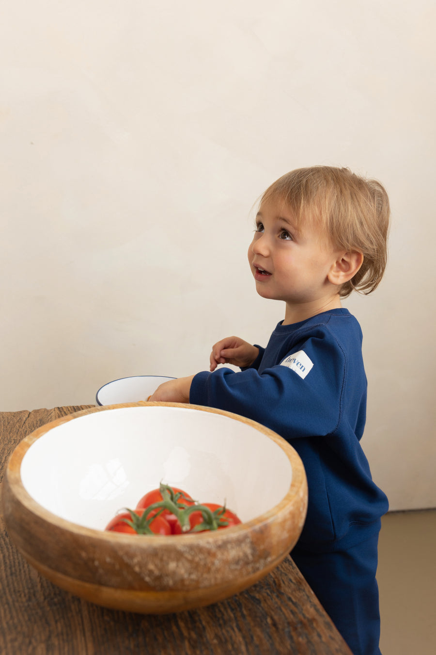 Child in a blue outfit standing next to a wooden bowl with tomatoes on a wooden table.