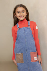 Young girl wearing a red long-sleeve shirt and blue denim overall dress against a beige background