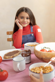 Young girl sitting at a table with food, including a bowl of cereal and a jar of peanut butter.