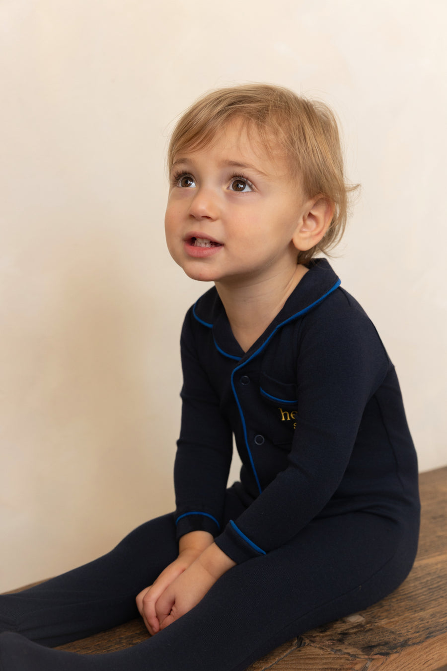 Child wearing a navy blue outfit with a collar, sitting on a wooden floor against a beige wall.