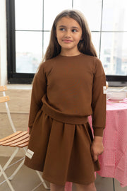 Young girl wearing a brown dress standing indoors with a checkered tablecloth and chair in the background.