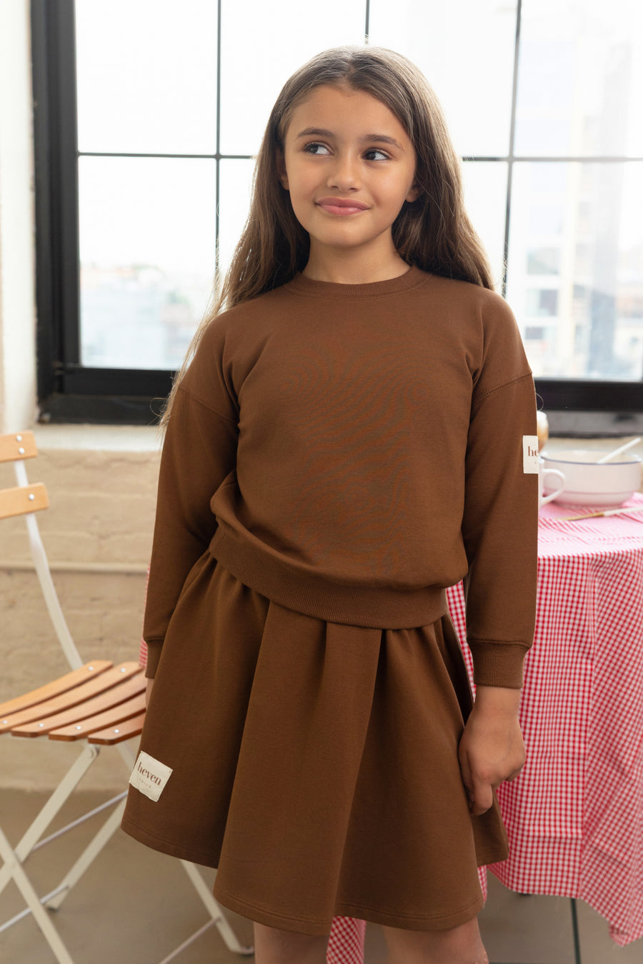 Young girl wearing a brown dress standing indoors with a checkered tablecloth and chair in the background.