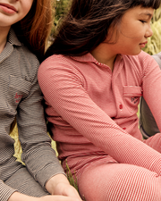 Two girls wearing striped long-sleeve polo shirts and matching pants, sitting outdoors on grass. The girl on the right wears a pink and white striped shirt, while the girl on the left wears a gray and white striped shirt.