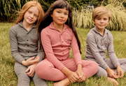 Three children sitting on grass outdoors wearing matching long-sleeve pajama sets in gray, pink, and light gray.